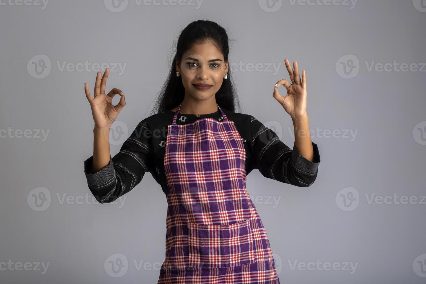 Portrait of Indian woman chef or cook in an apron, presenting, pointing, with ok sign, thumbs up on the grey background. photo