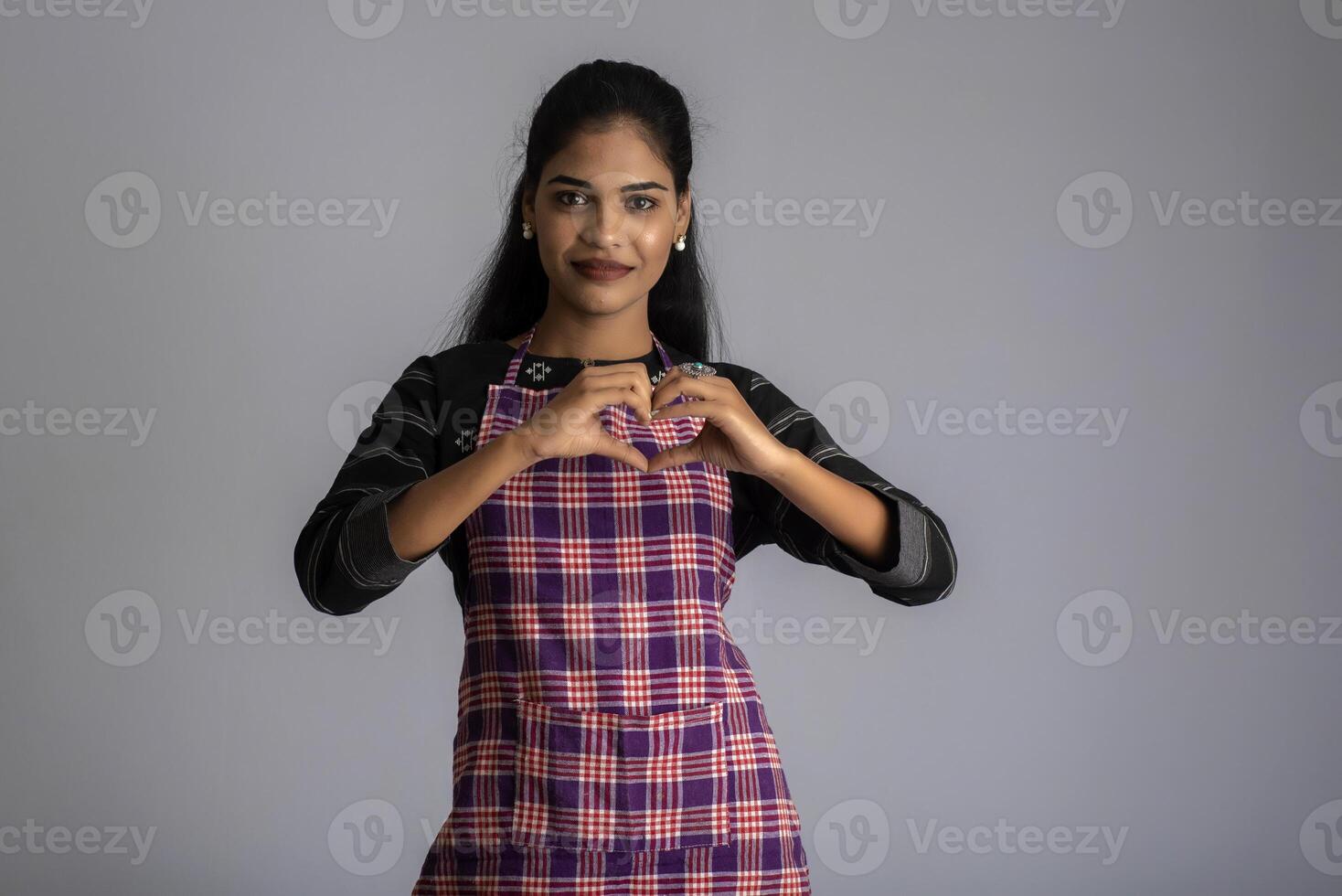 Portrait of Indian woman chef or cook in an apron, presenting, pointing, with ok sign, thumbs up on the grey background. photo