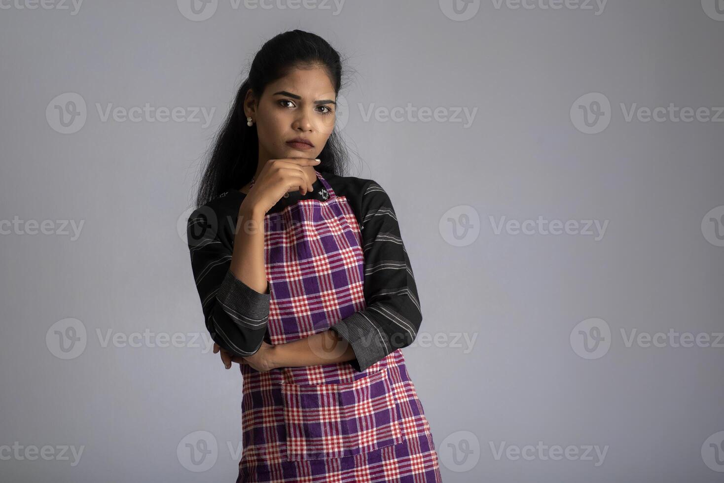 Portrait of Indian woman chef or cook in an apron, presenting, pointing, with ok sign, thumbs up on the grey background. photo
