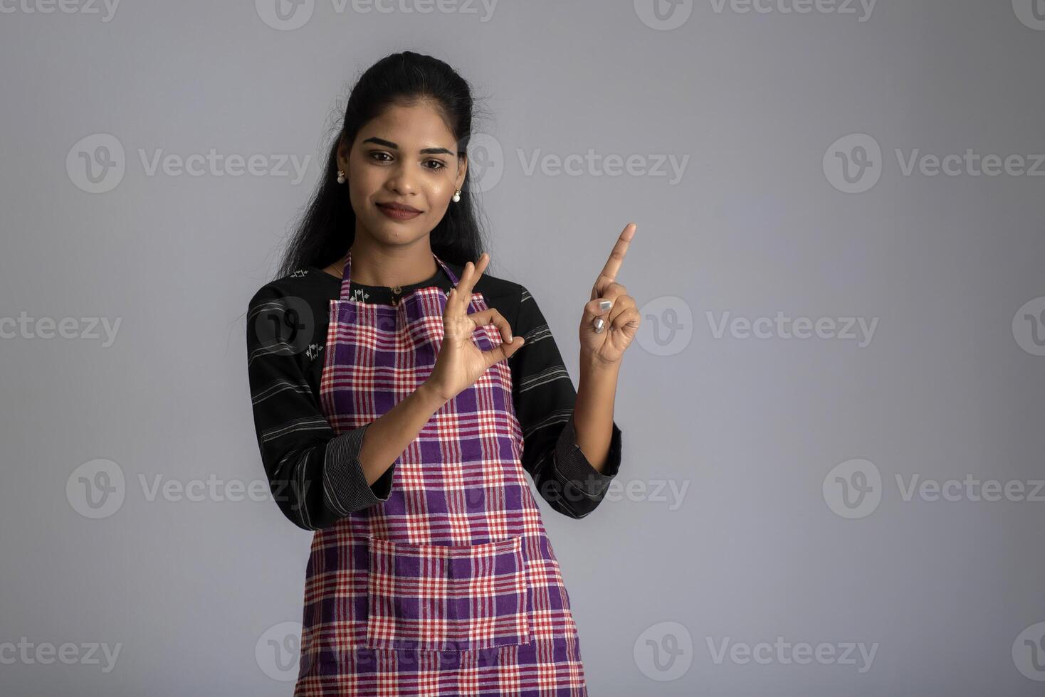 Portrait of Indian woman chef or cook in an apron, presenting, pointing, with ok sign, thumbs up on the grey background. photo