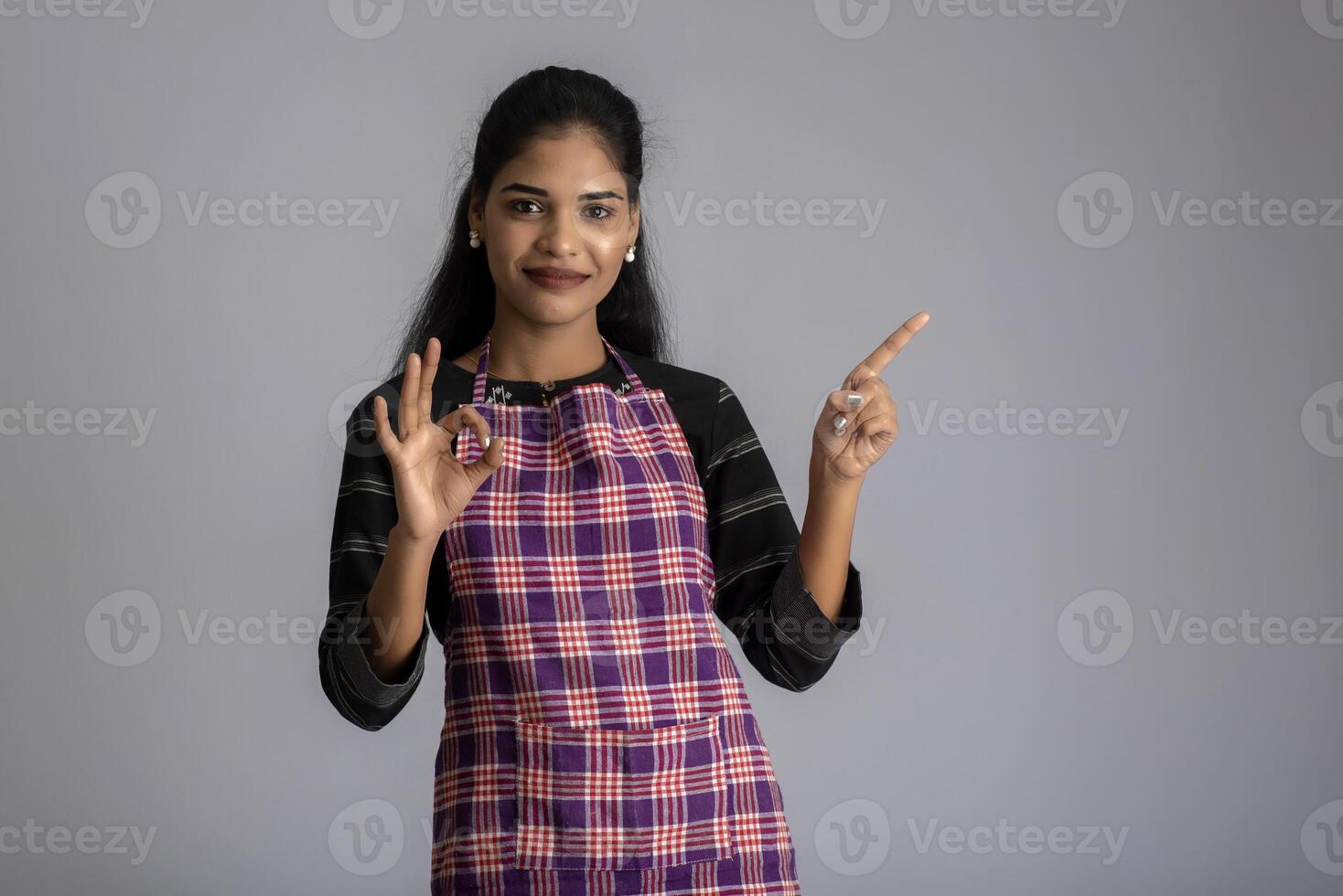 Portrait of Indian woman chef or cook in an apron, presenting, pointing, with ok sign, thumbs up on the grey background. photo
