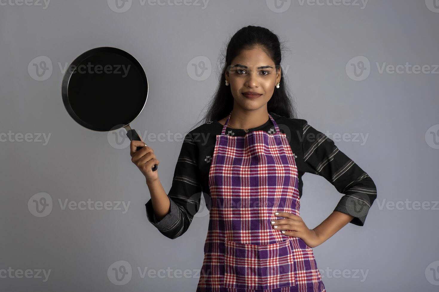 Young girl holding kitchen utensils spatula and pan on a grey background photo