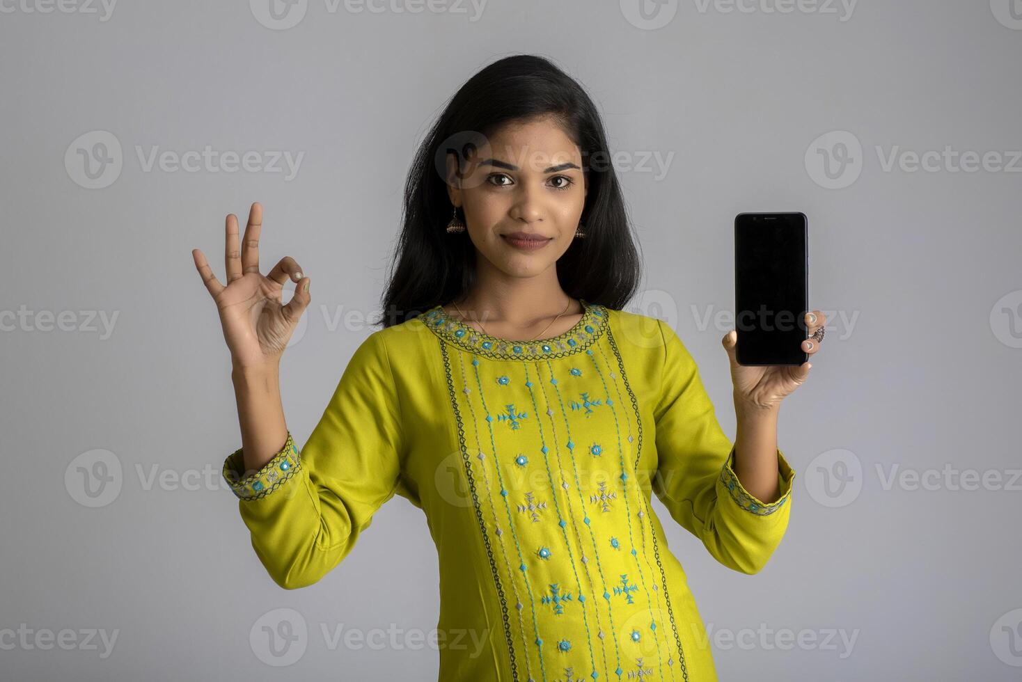 Young beautiful girl holding and showing blank screen of smartphone or mobile or tablet phone on a gray background. photo