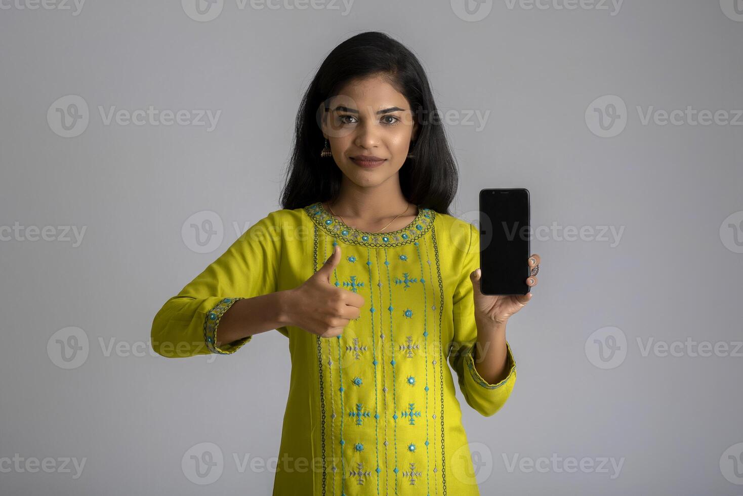 Young beautiful girl holding and showing blank screen of smartphone or mobile or tablet phone on a gray background. photo