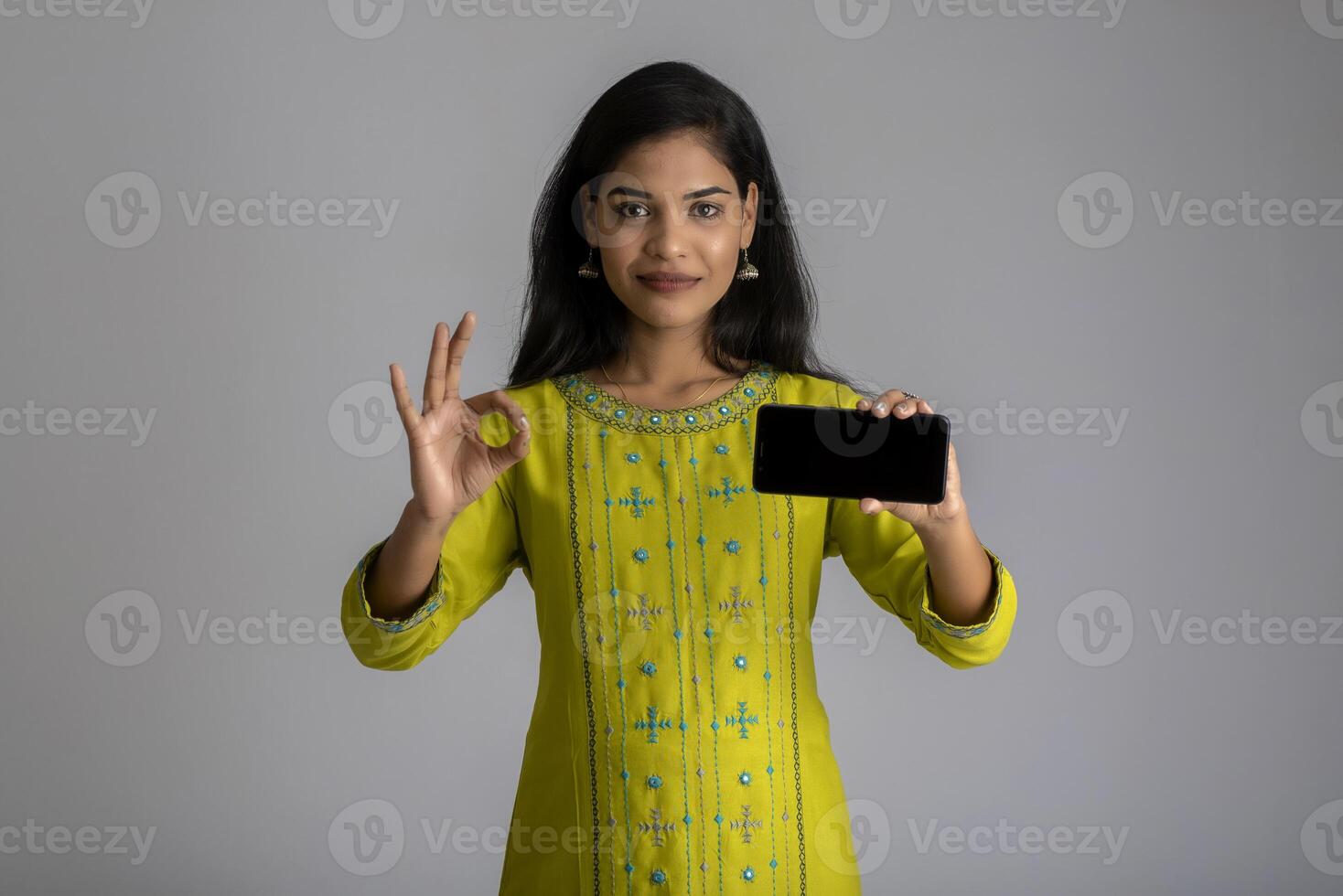 Young beautiful girl holding and showing blank screen of smartphone or mobile or tablet phone on a gray background. photo