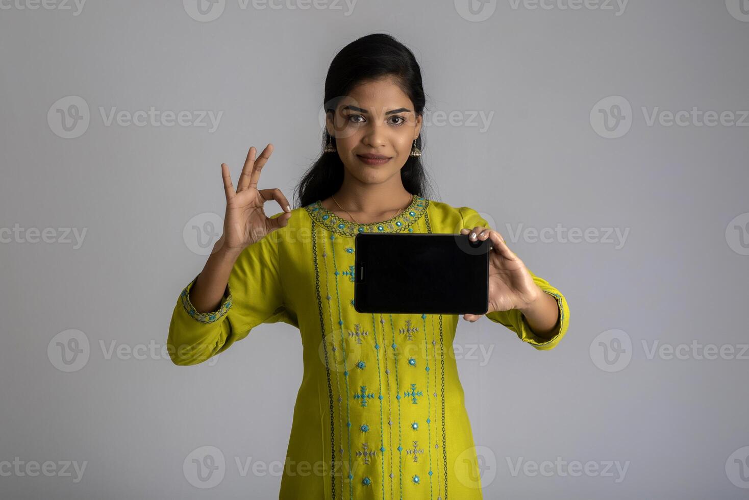 Young beautiful girl holding and showing blank screen of smartphone or mobile or tablet phone on a gray background. photo