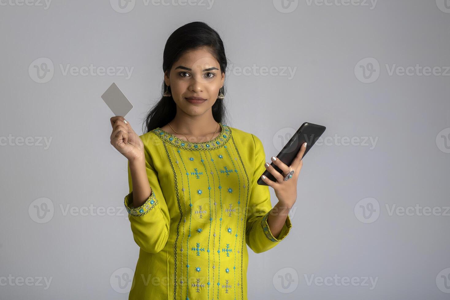 Young smiling beautiful woman or girl presenting credit card while using mobile or smartphone on grey background. photo