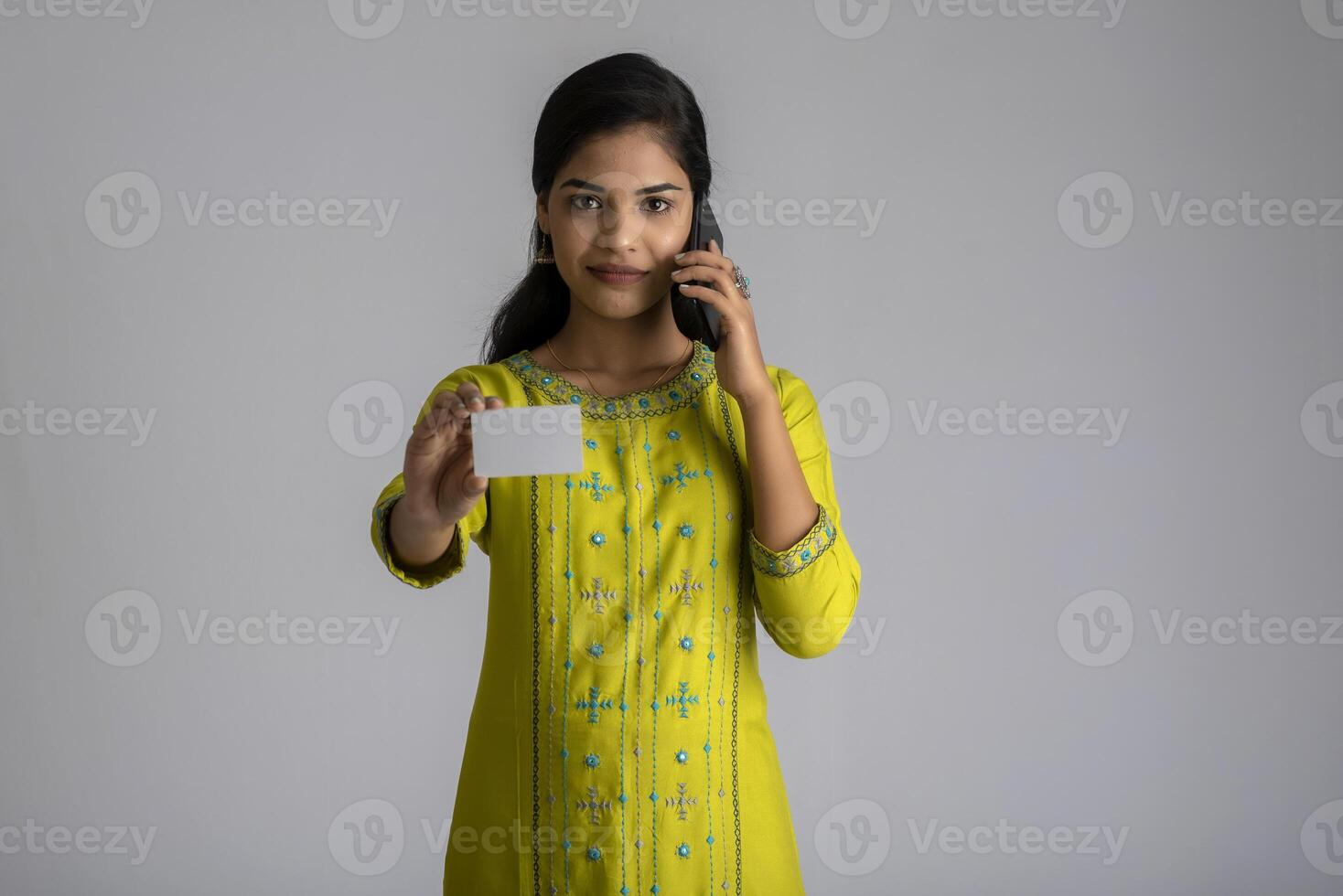 Young smiling beautiful woman or girl presenting credit card while using mobile or smartphone on grey background. photo