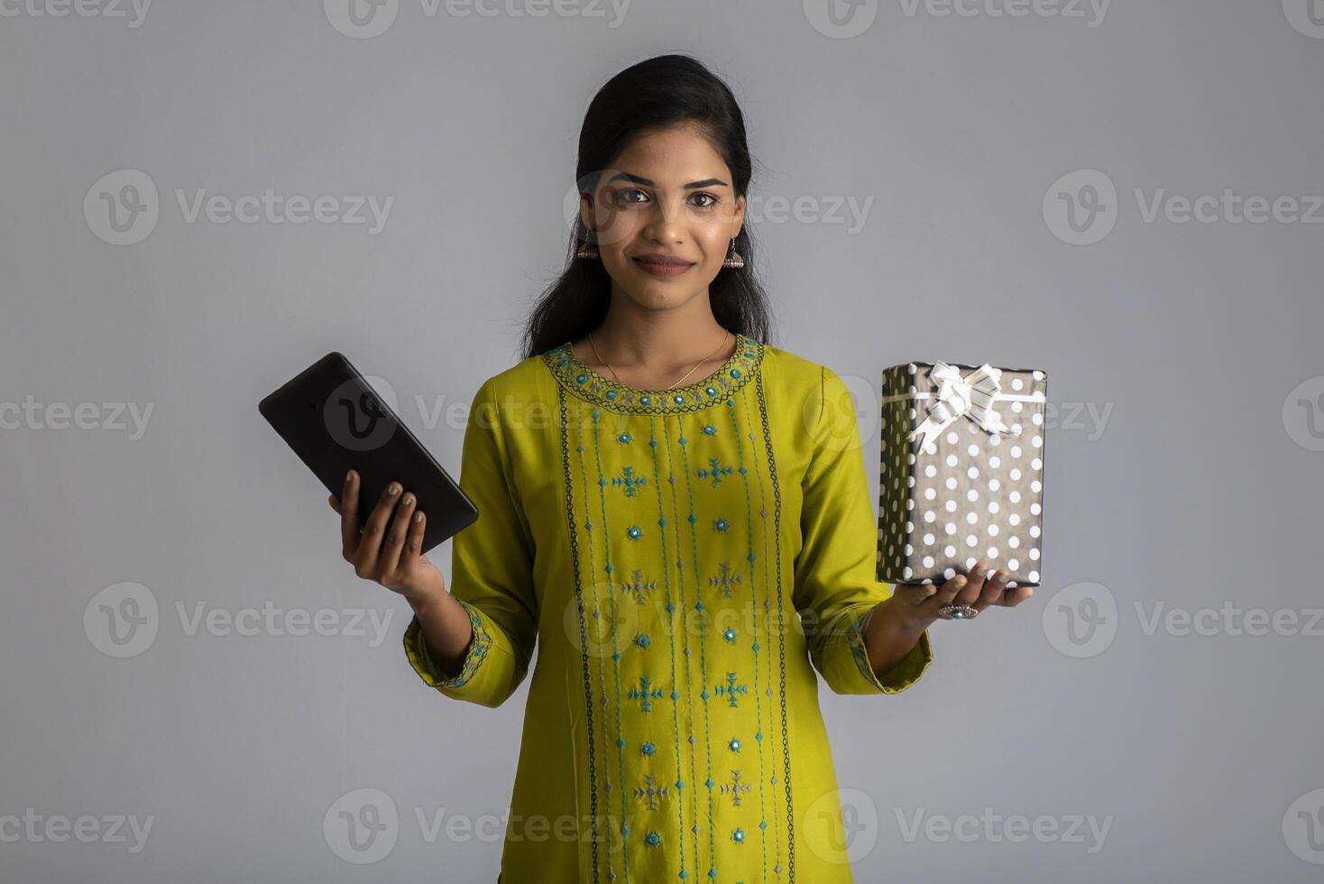 Portrait of young happy smiling Girl holding gift box and showing smartphone or tablet on a grey background. photo