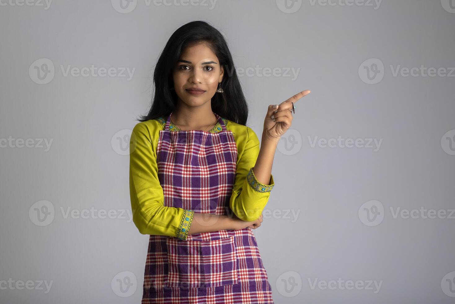 Portrait of Indian woman chef or cook in an apron, presenting, pointing, with ok sign, thumbs up on the grey background. photo