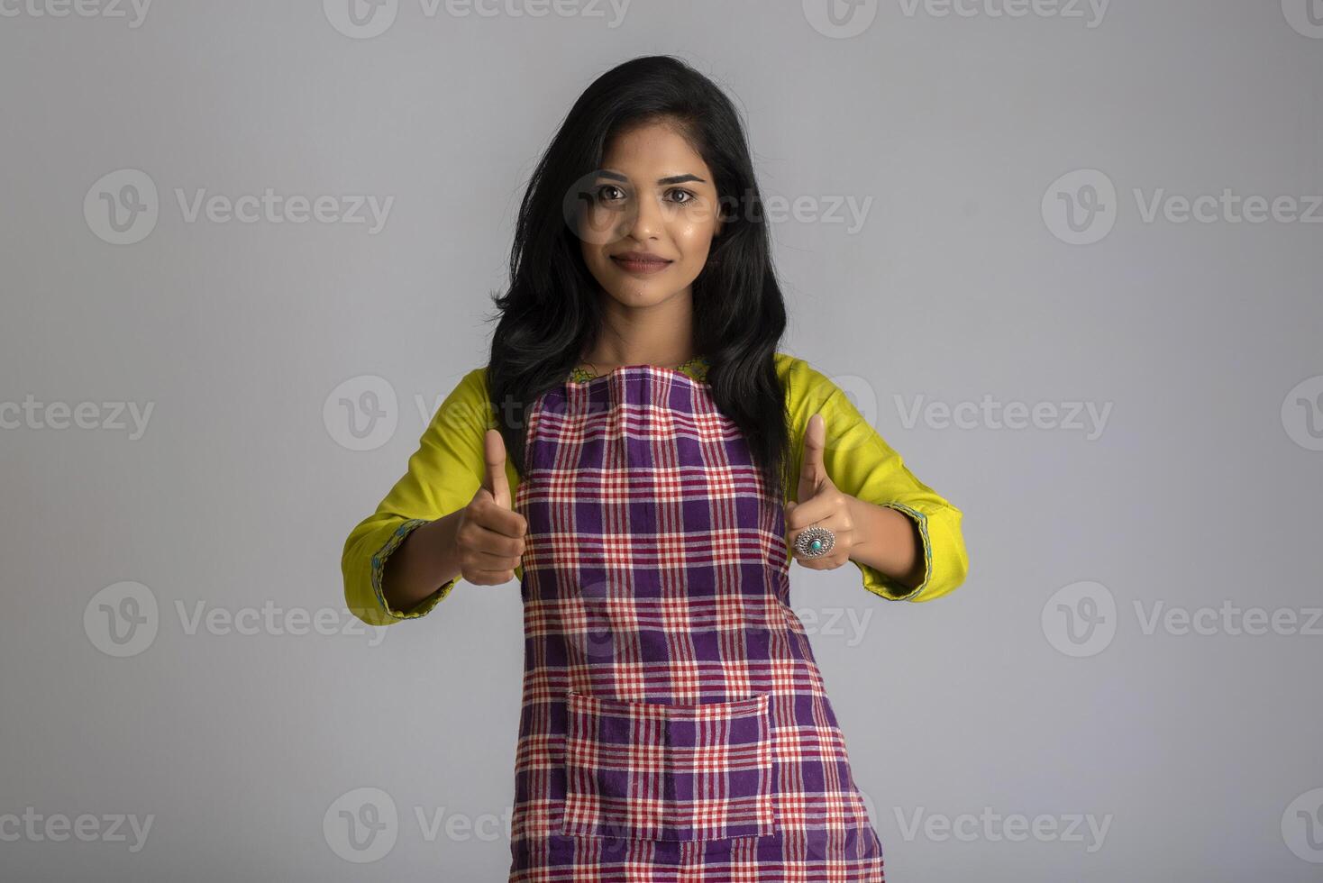 Portrait of Indian woman chef or cook in an apron, presenting, pointing, with ok sign, thumbs up on the grey background. photo