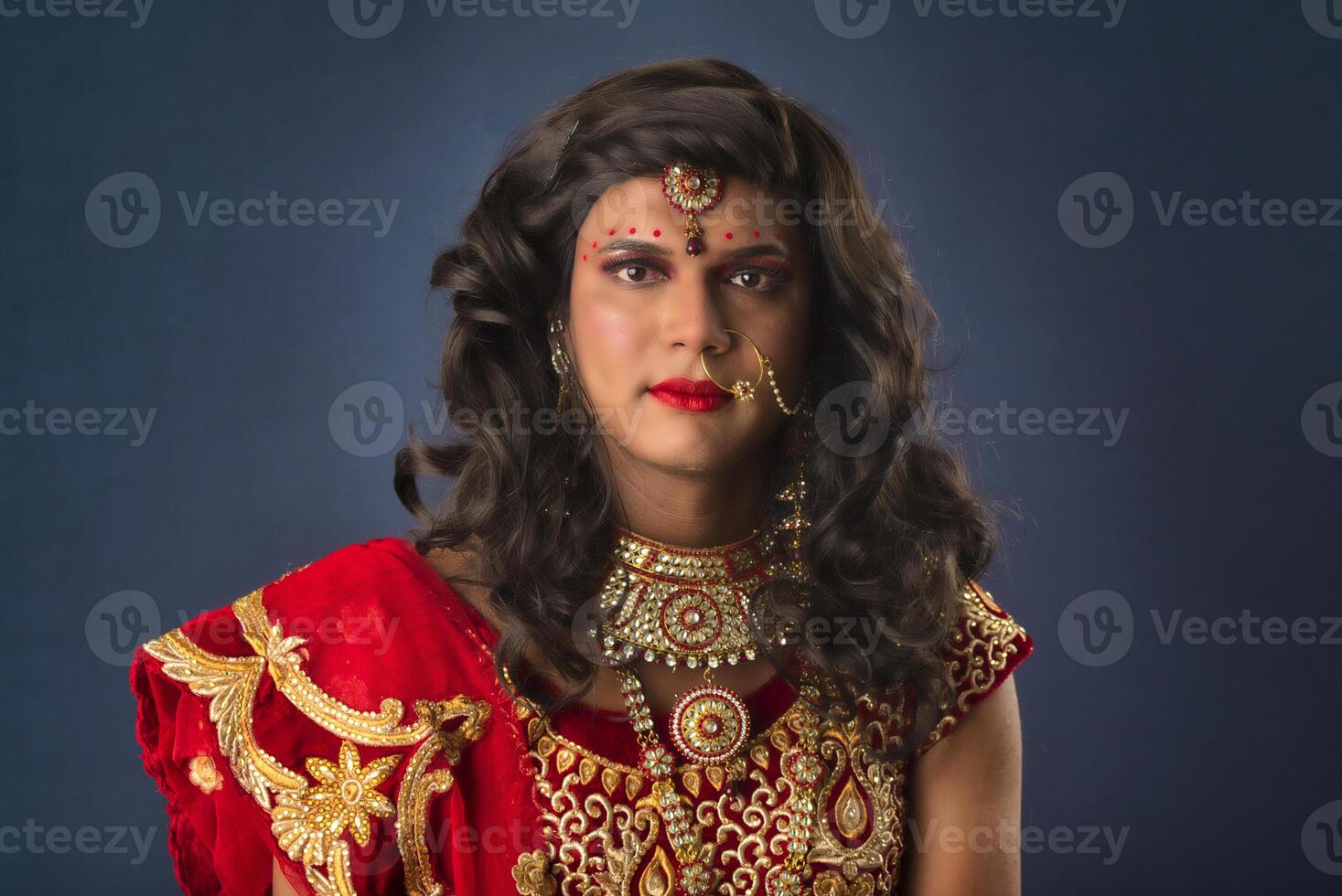 Young crossdresser man wearing Indian wedding clothes of a bride with bridal makeup and posing on grey background, looking fashion and glamorous. photo