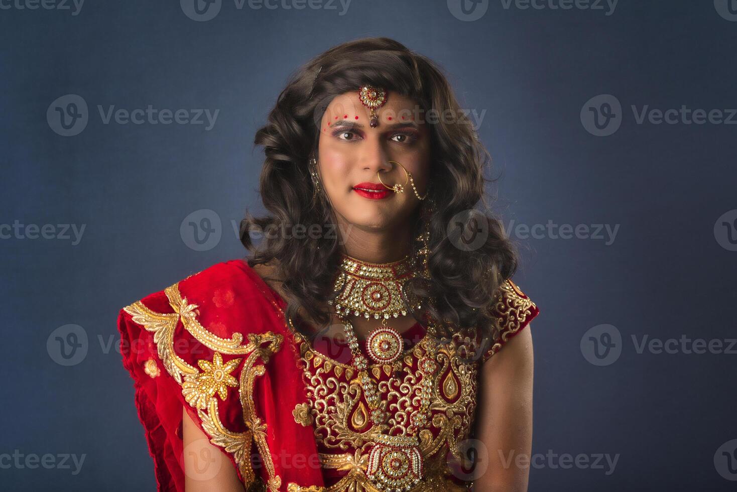 Young crossdresser man wearing Indian wedding clothes of a bride with bridal makeup and posing on grey background, looking fashion and glamorous. photo
