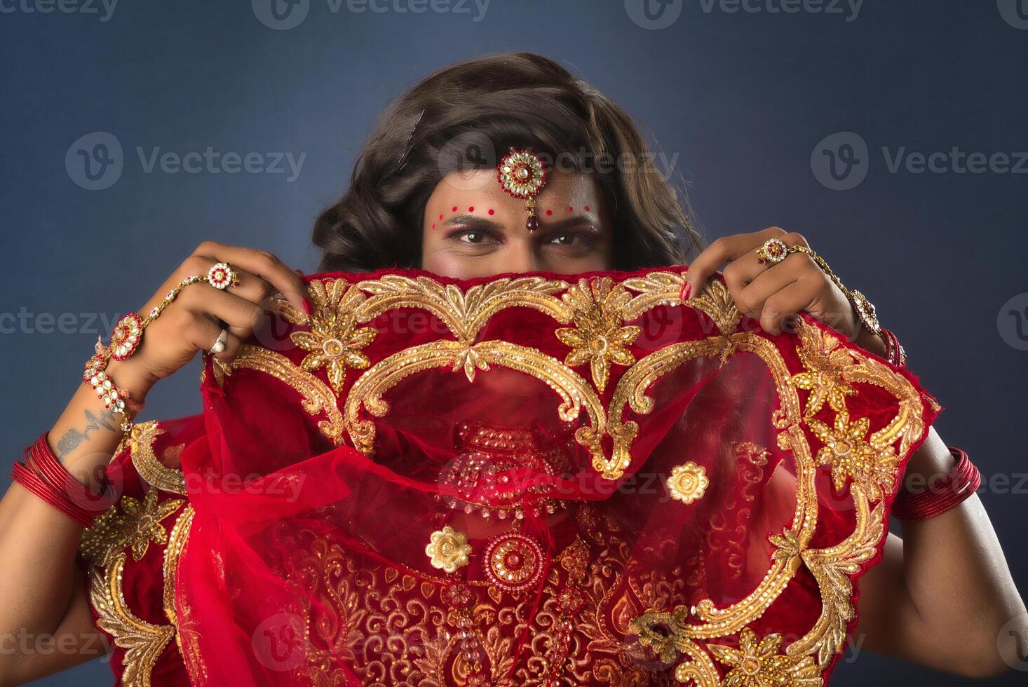 Young crossdresser man wearing Indian wedding clothes of a bride with bridal makeup and posing on grey background, looking fashion and glamorous. photo