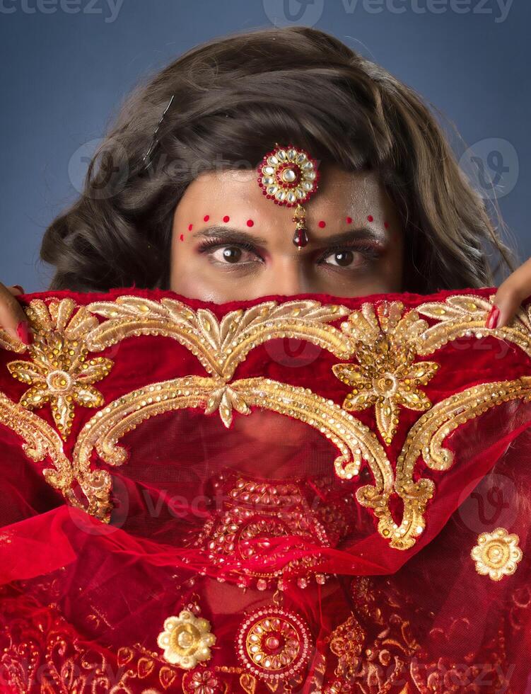 Young crossdresser man wearing Indian wedding clothes of a bride with bridal makeup and posing on grey background, looking fashion and glamorous. photo