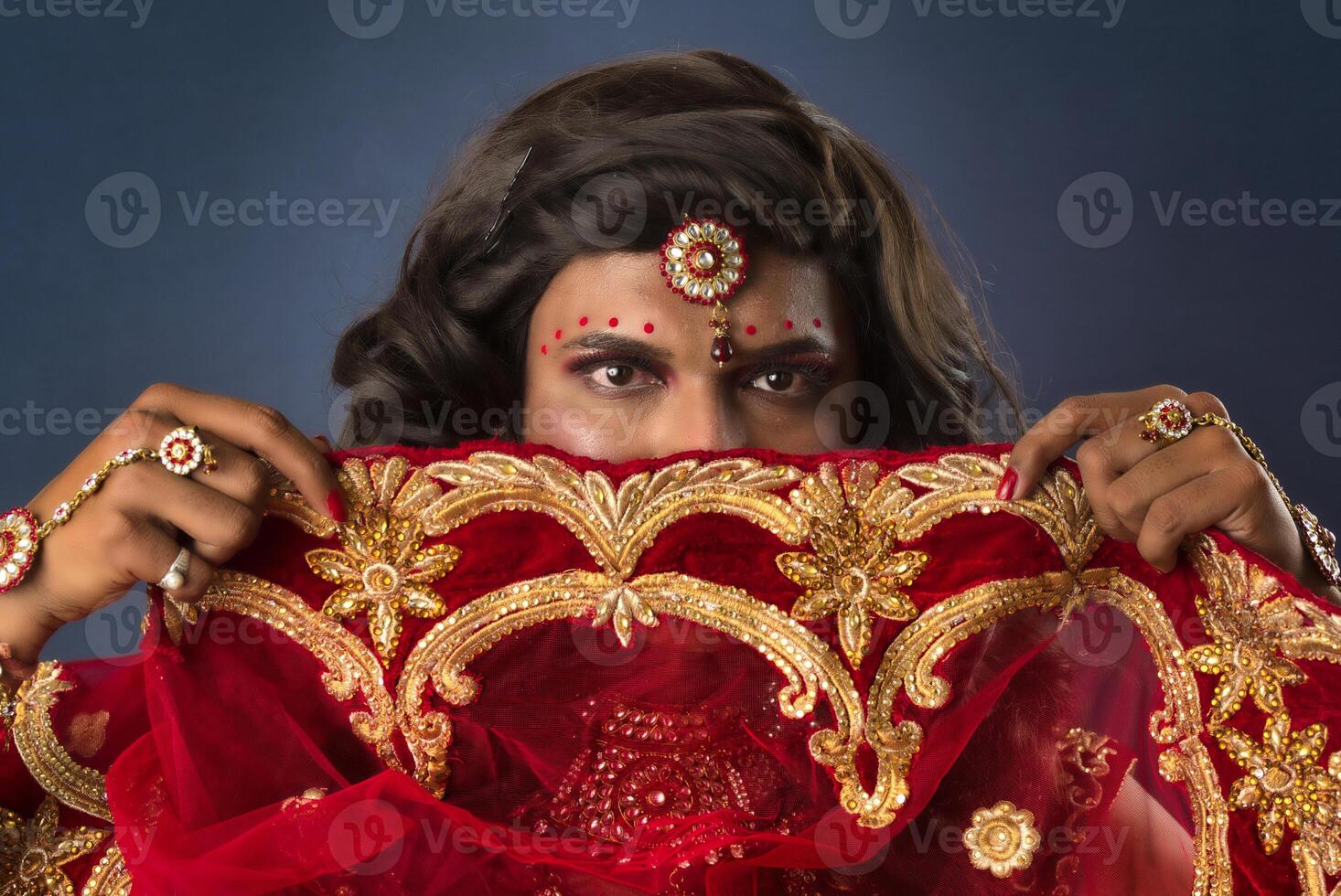 Young crossdresser man wearing Indian wedding clothes of a bride with bridal makeup and posing on grey background, looking fashion and glamorous. photo