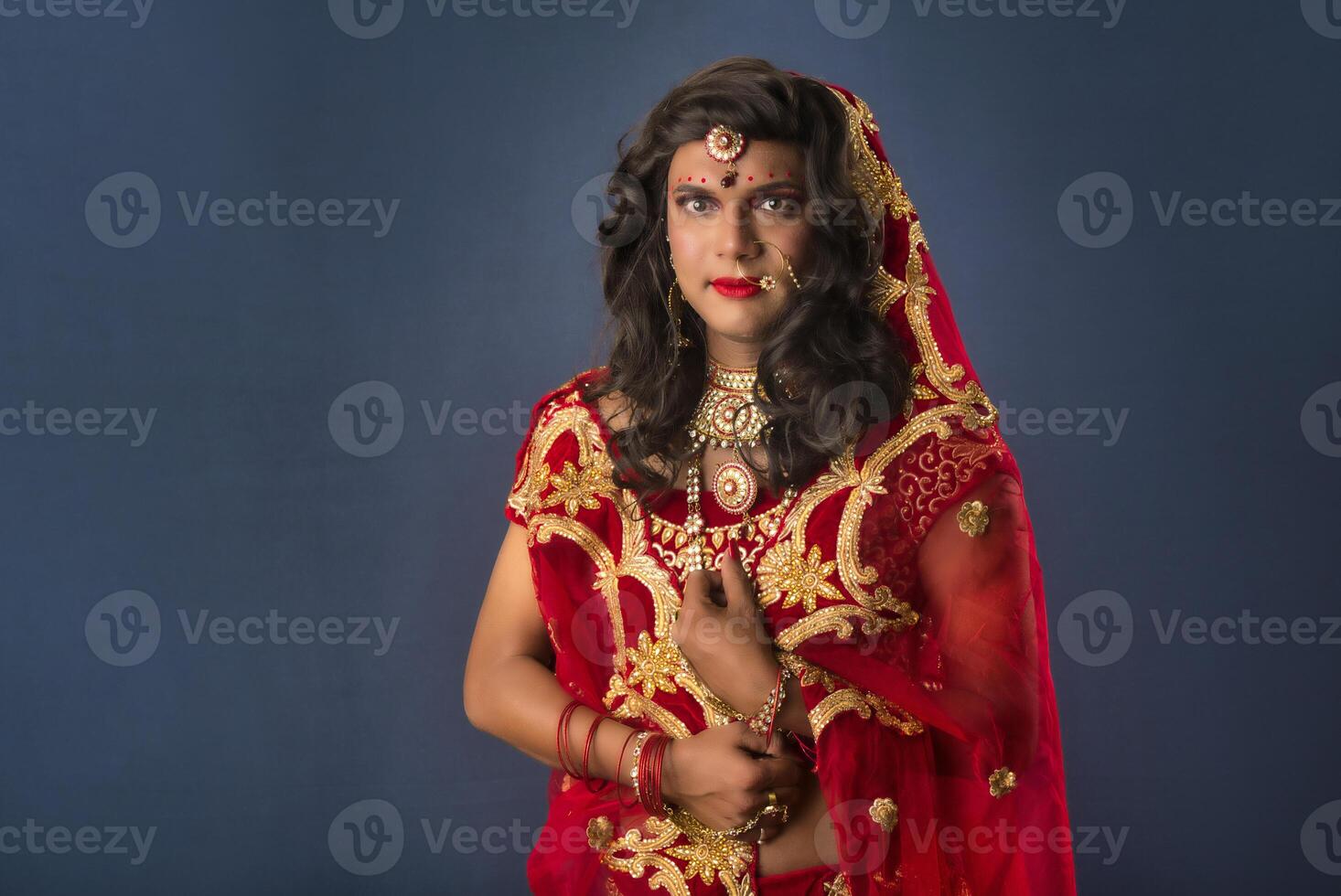 Young crossdresser man wearing Indian wedding clothes of a bride with bridal makeup and posing on grey background, looking fashion and glamorous. photo