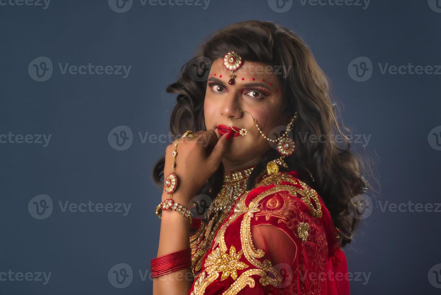 Young crossdresser man wearing Indian wedding clothes of a bride with bridal makeup and posing on grey background, looking fashion and glamorous. photo
