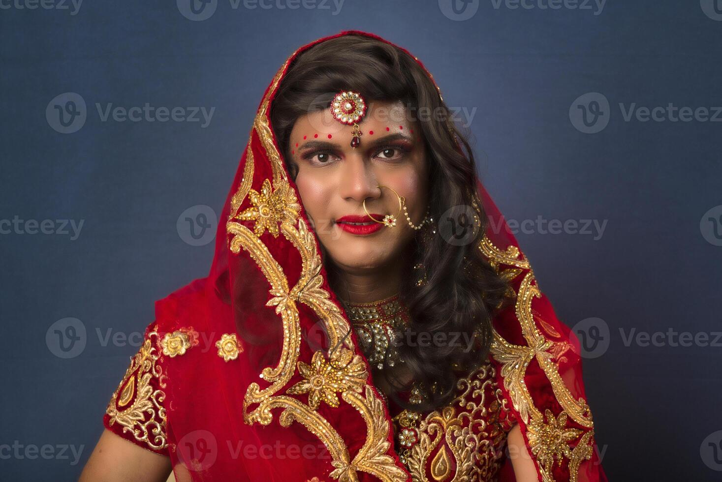 Young crossdresser man wearing Indian wedding clothes of a bride with bridal makeup and posing on grey background, looking fashion and glamorous. photo