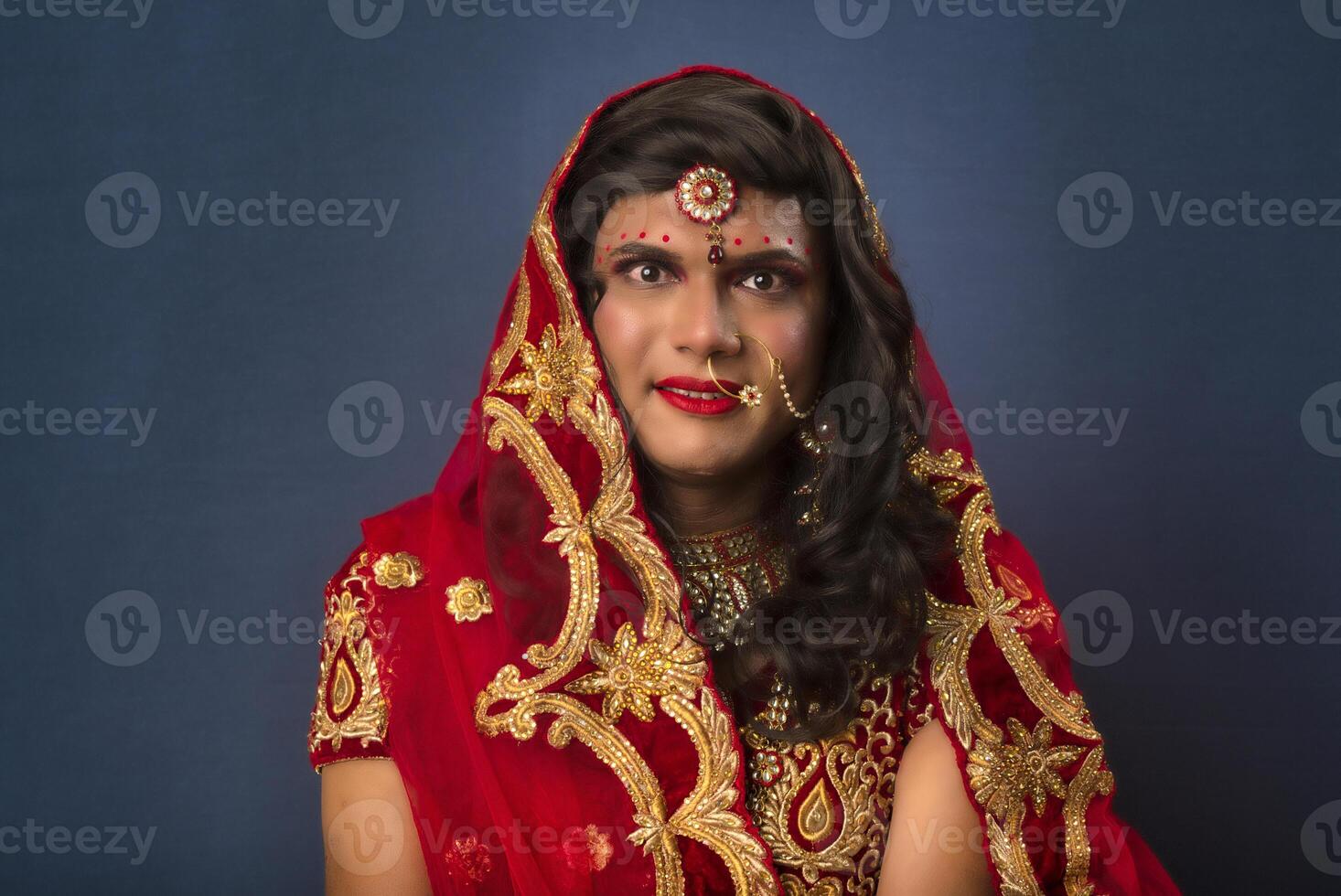 Young crossdresser man wearing Indian wedding clothes of a bride with bridal makeup and posing on grey background, looking fashion and glamorous. photo