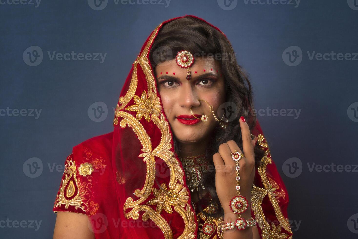 Young crossdresser man wearing Indian wedding clothes of a bride with bridal makeup and posing on grey background, looking fashion and glamorous. photo