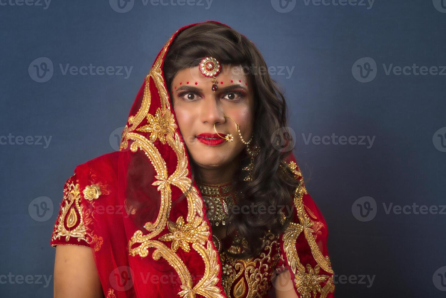 Young crossdresser man wearing Indian wedding clothes of a bride with bridal makeup and posing on grey background, looking fashion and glamorous. photo