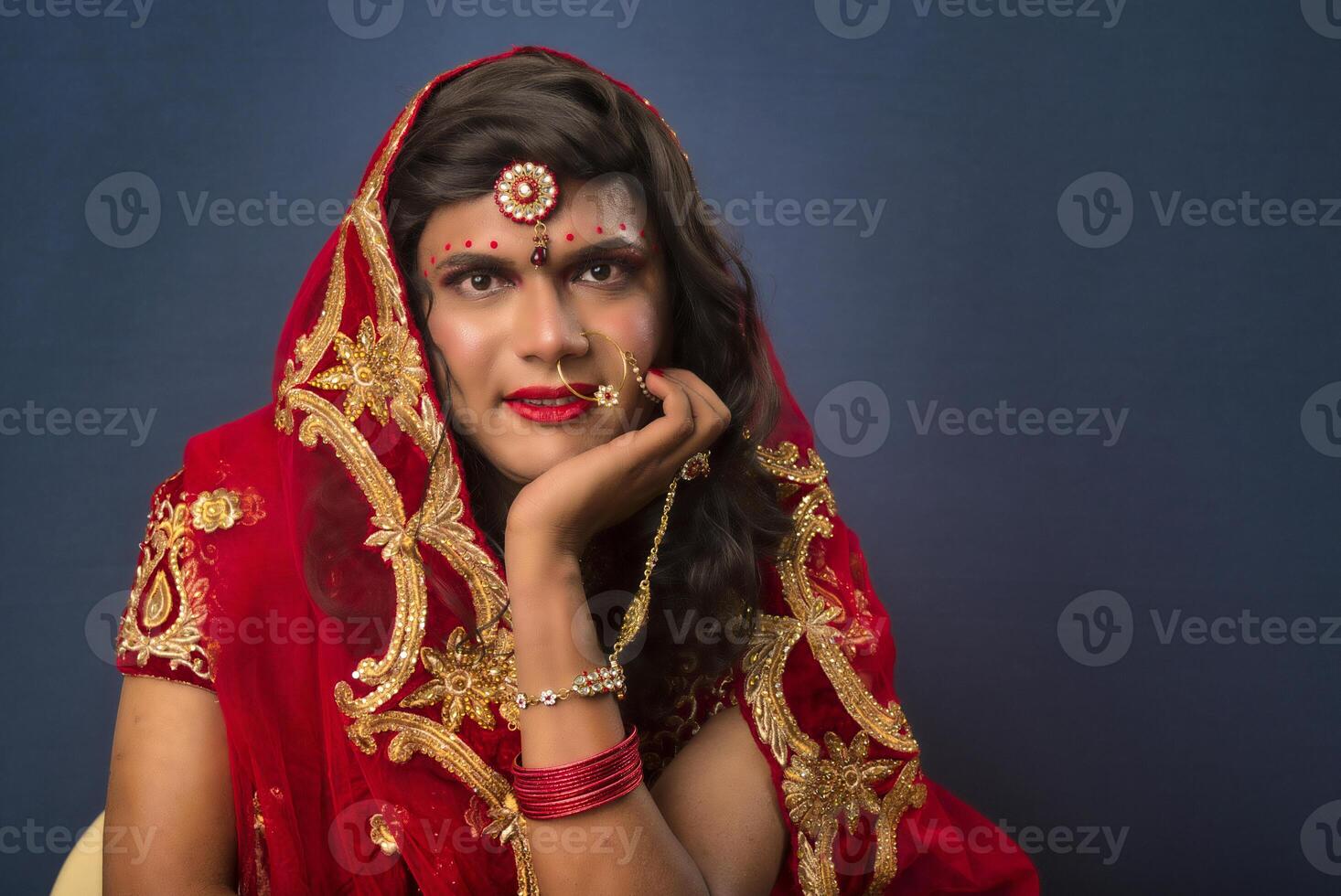 Young crossdresser man wearing Indian wedding clothes of a bride with bridal makeup and posing on grey background, looking fashion and glamorous. photo