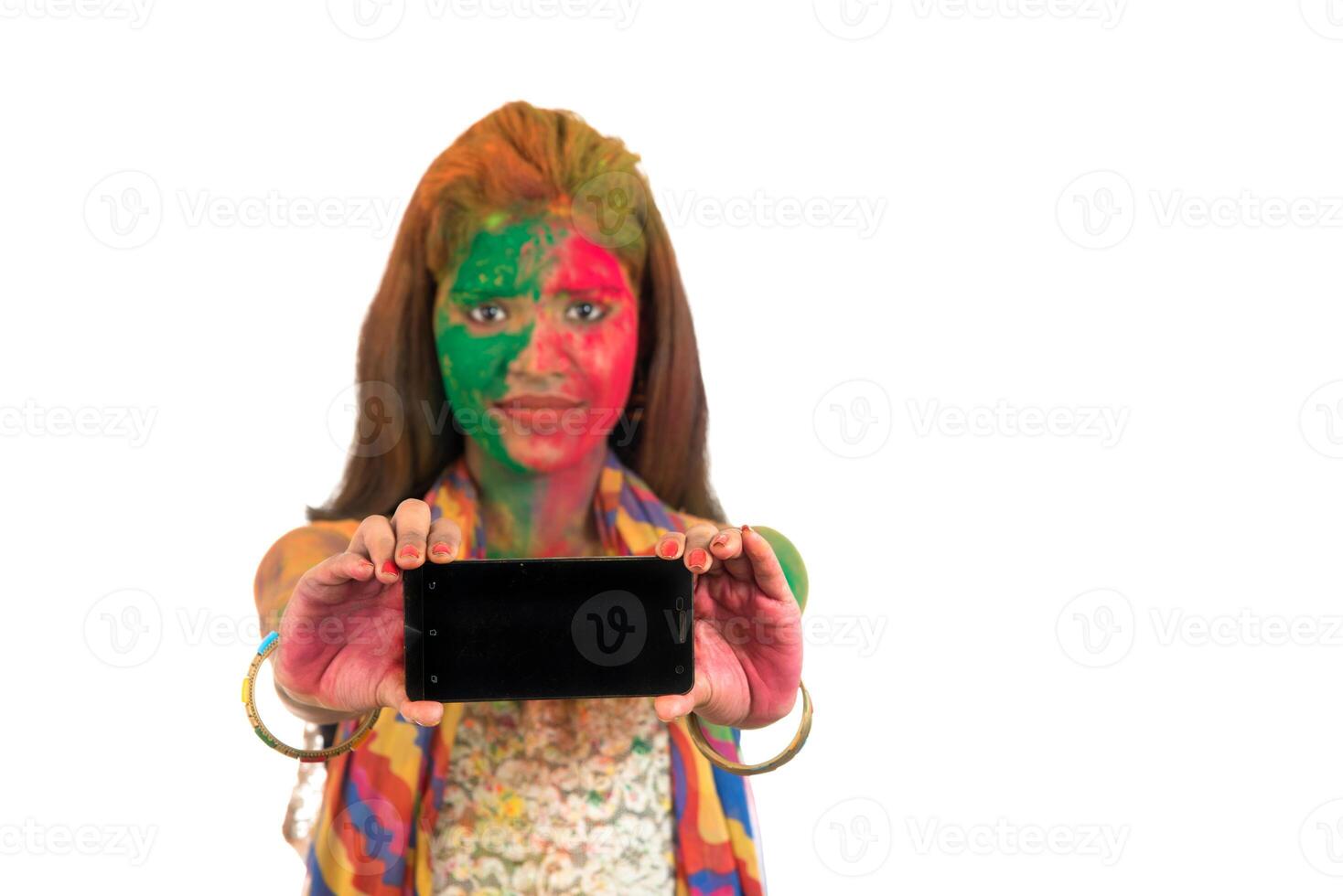 Young girl with colorful face showing smart phone screen on Holi festival. Festival and technology concept on white background. photo