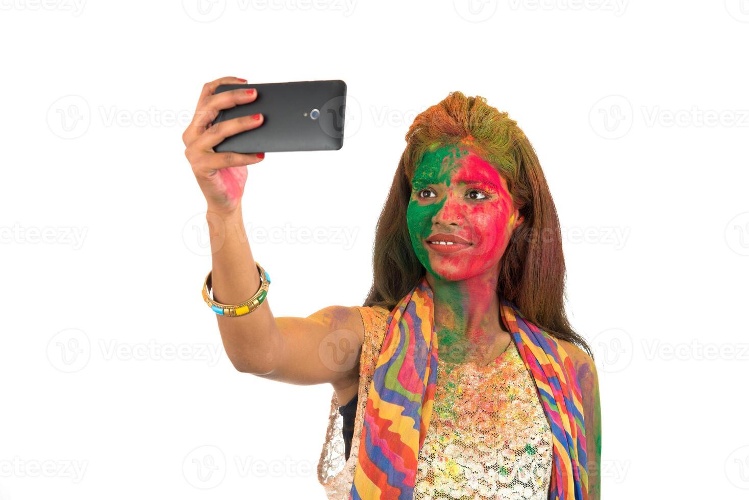 Young girl with colourful face taking selfie using smartphone on Holi festival. Festival and technology concept on white background. photo