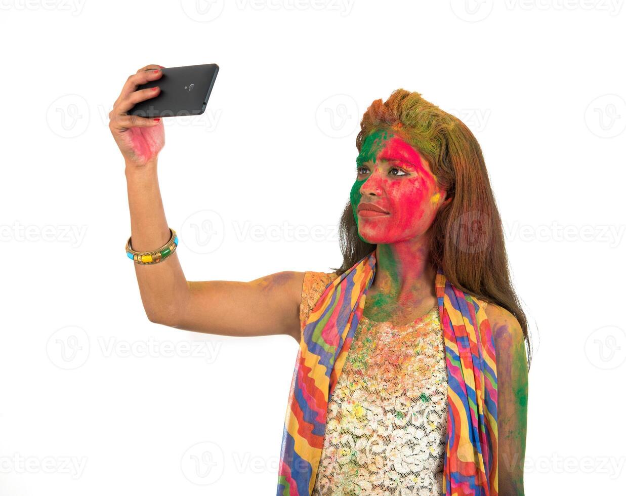 Young girl with colourful face taking selfie using smartphone on Holi festival. Festival and technology concept on white background. photo