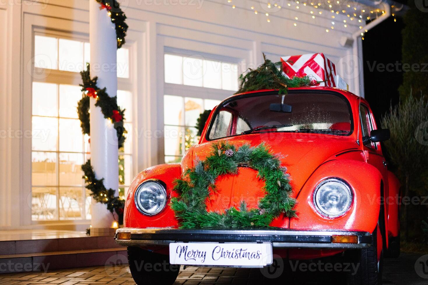 A red retro car decorated for Christmas with gifts on the roof stands near the house in the evening photo