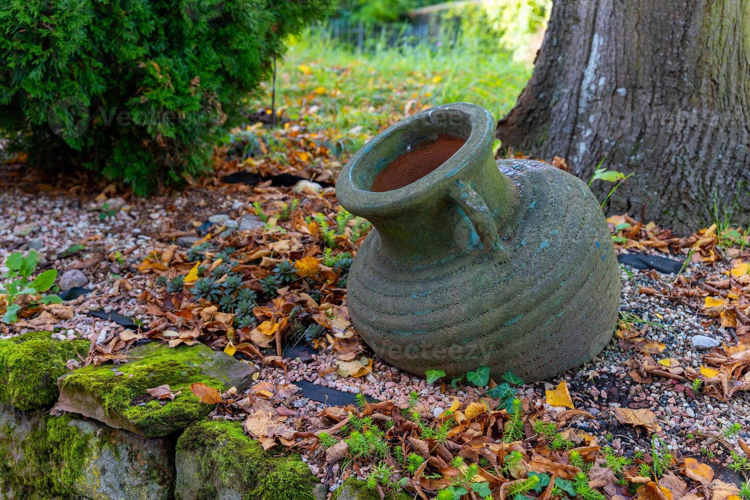 A large, green clay pot sits beneath a tree in a garden with fallen leaves photo