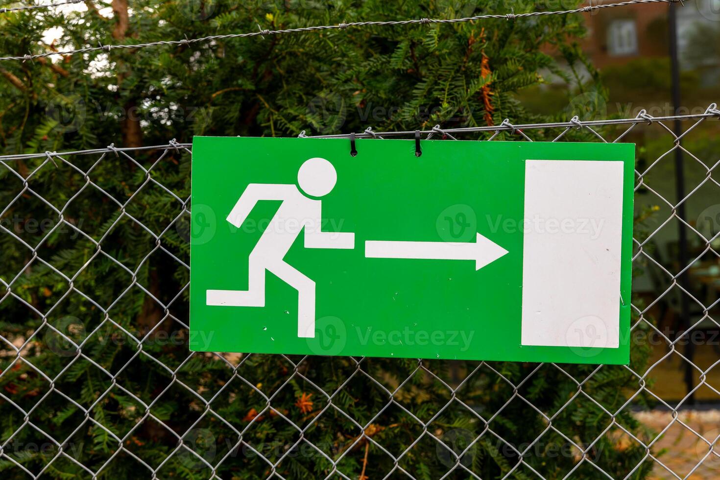 A green exit sign with a white stick figure running right is attached to a chain-link fence photo