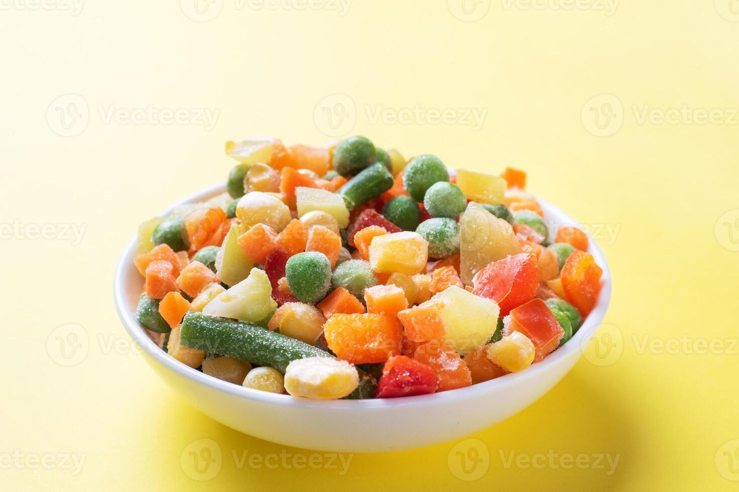 Chopped frozen vegetables in a plate on a yellow background. Corn peas pepper carrots. copy space. photo