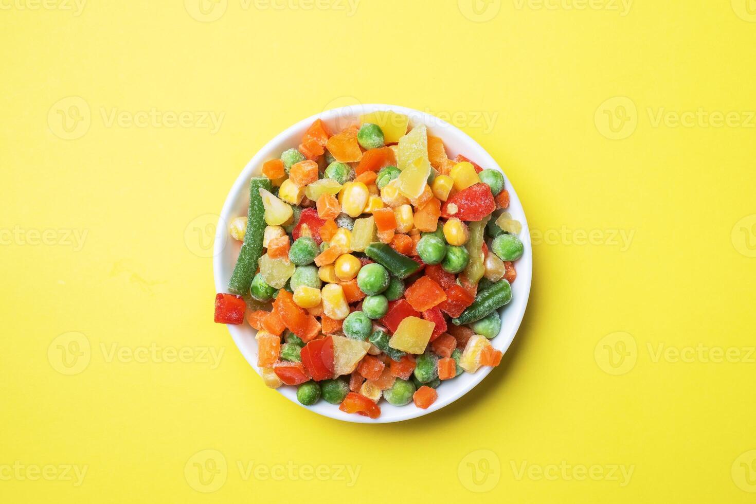 Chopped frozen vegetables in a plate on a yellow background. Corn peas pepper carrots. copy space. photo