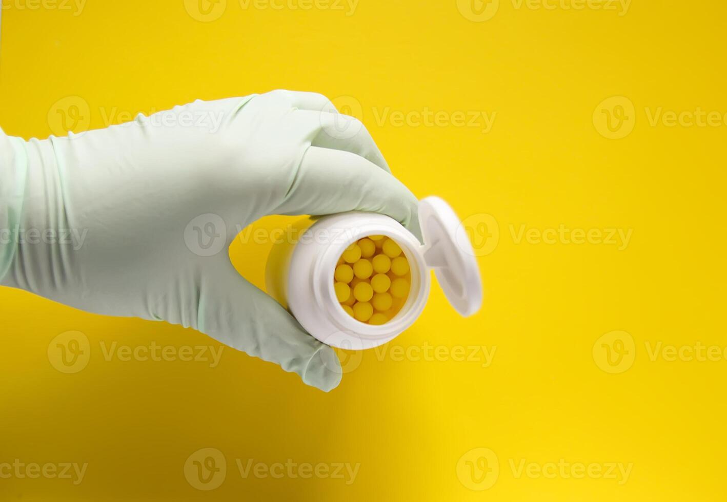 Hand in latex glove holding the plastic container of pills on bright yellow background. photo