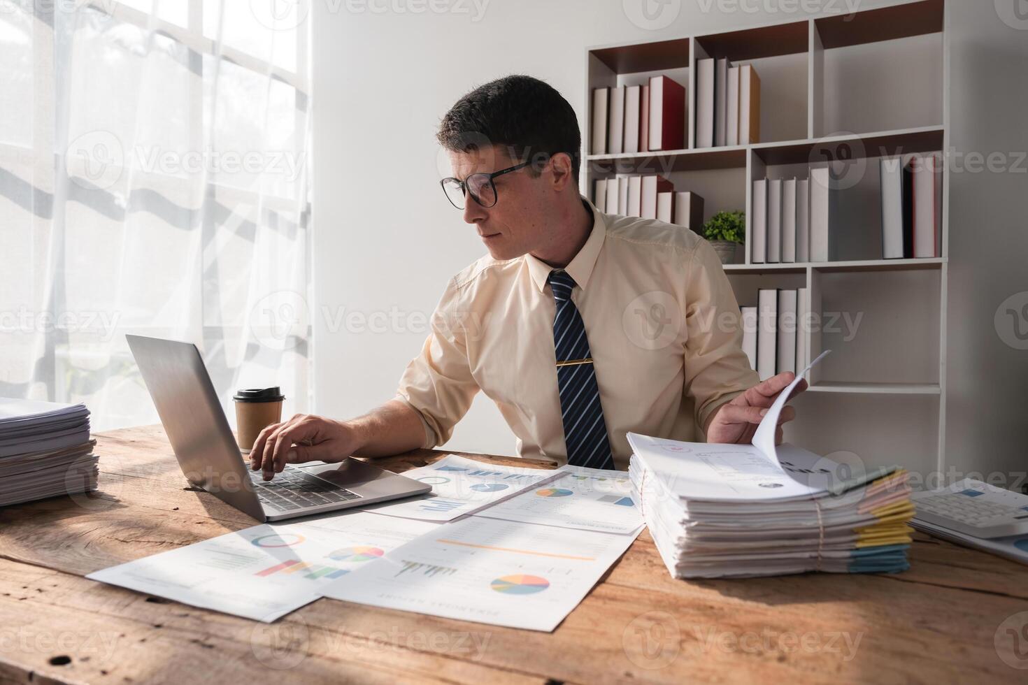Businessman Working in Modern Office with Documents and Laptop, Analyzing Data and Reports, Professional Workspace, Daylight through Windows, Focused and Productive Work Environment photo