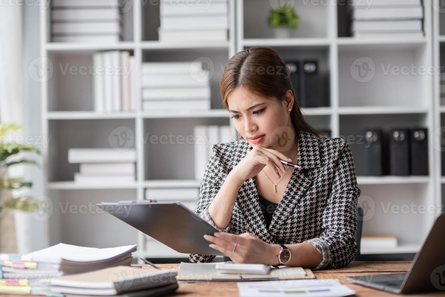 Young woman stressed at work, sitting at a desk with documents, holding a tablet, looking worried and overwhelmed in a modern office environment with shelves and plants in the background. photo