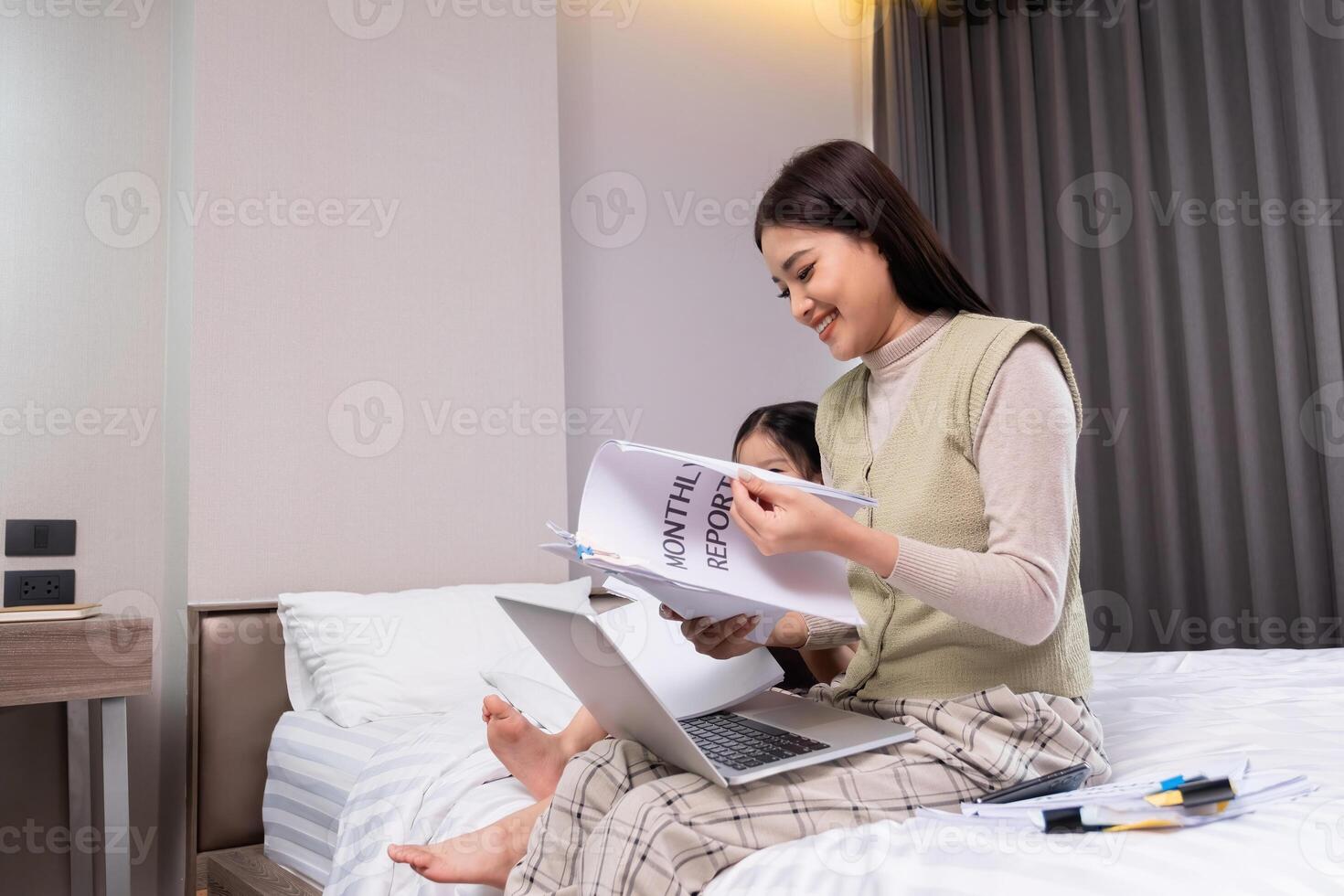 Middle-aged woman working from home, sitting on bed with laptop and documents, multitasking in a modern bedroom setting, showcasing remote work lifestyle and home office environment. photo