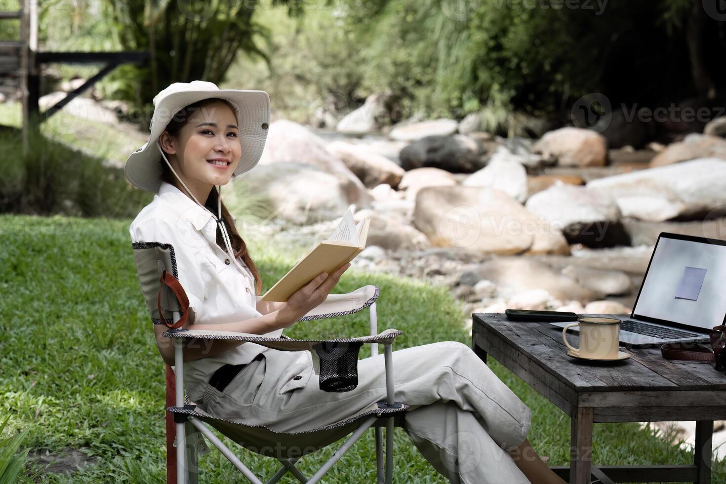 Young Woman Enjoying a Peaceful Camping Experience in the Forest with a Book and Laptop by a Stream photo
