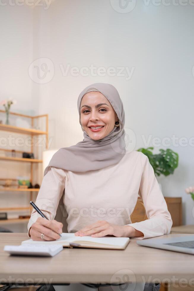 Confident Muslim Woman in Hijab Working at Desk, Smiling and Writing in Notebook, Modern Office Environment, Professional and Empowered, Diverse Workplace, Business and Education Concept photo