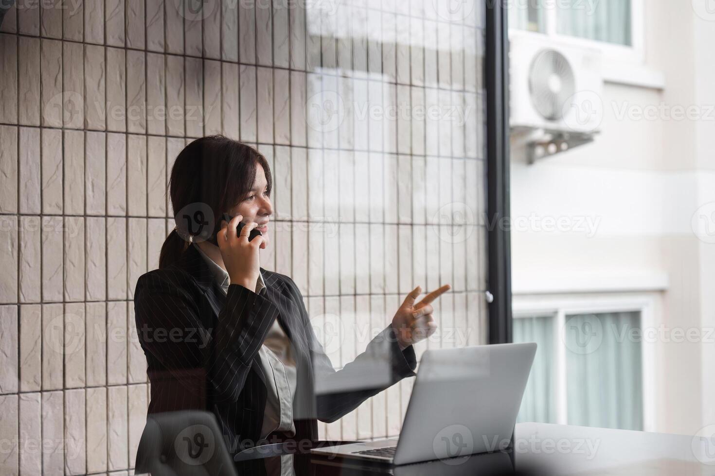 Professional Businesswoman Communicating with Clients via Telephone in Modern Office Setting with Laptop and Window View photo