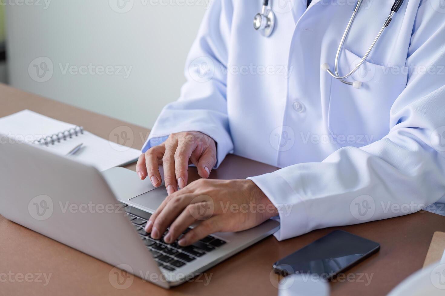 Male Doctor Using Laptop in Modern Healthcare Setting with Stethoscope and Smartphone on Desk photo