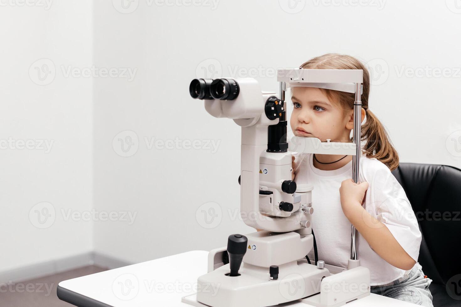 Portrait of a little, happy girl in ophthalmology clinic. Ophthalmic instrument. Excellent moment to illustrate careful examination for children's eye health. Advertisement for pediatric ophthalmology photo