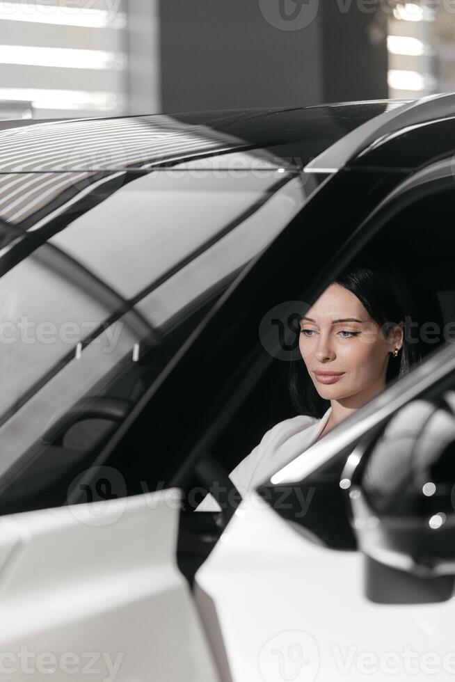 Portrait of gorgeous brunette woman make purchase in car showroom. Female sits inside of new, modern, electric car and look at sales agent. Concept for car rental or sale. Test drive. photo