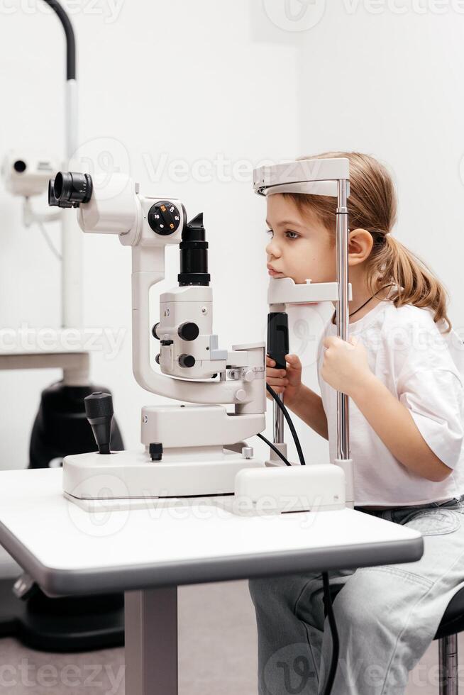 Portrait of a little, happy girl in ophthalmology clinic. Ophthalmic instrument. Excellent moment to illustrate careful examination for children's eye health. Advertisement for pediatric ophthalmology photo
