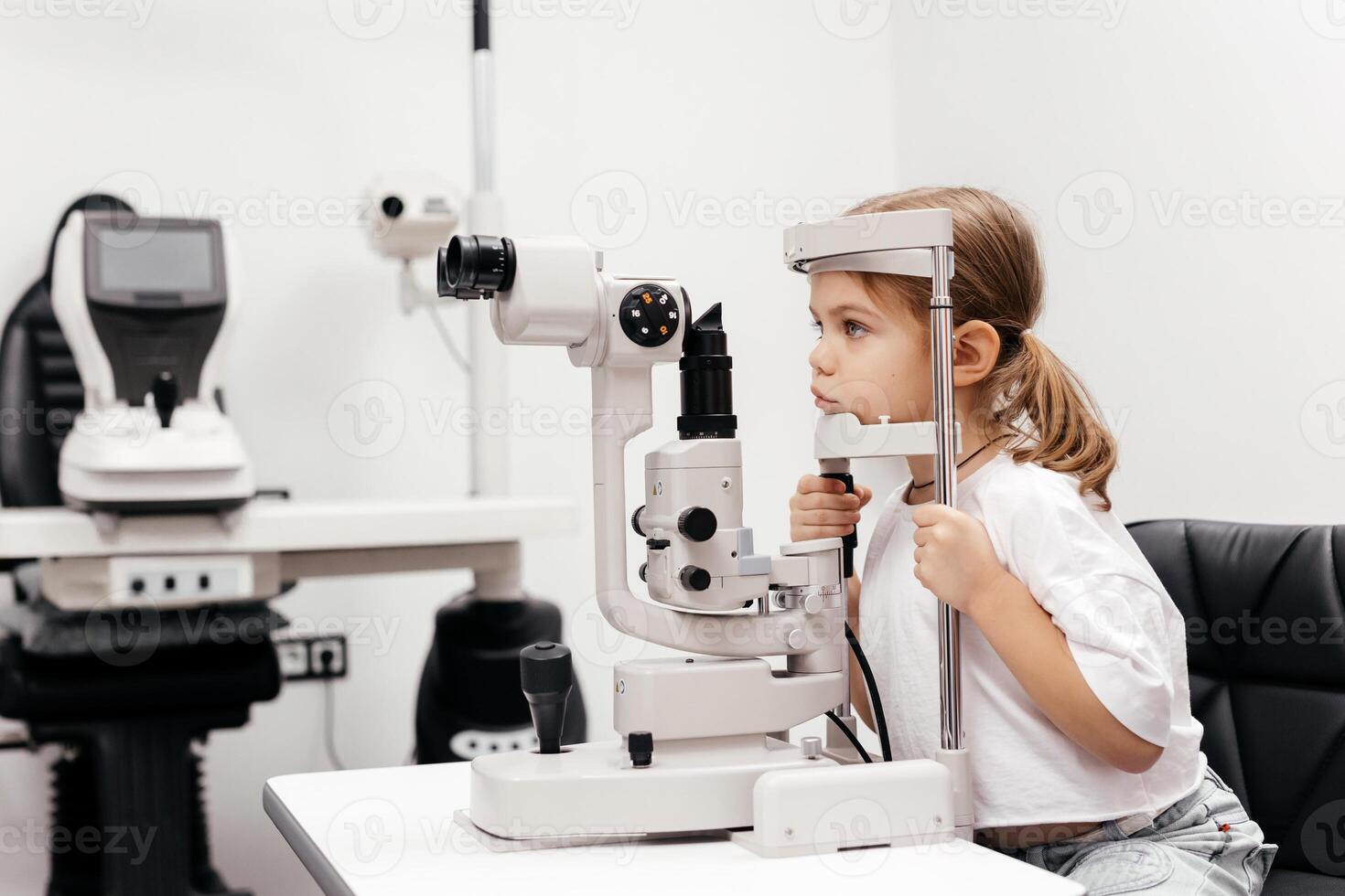 Portrait of a little, happy girl in ophthalmology clinic. Ophthalmic instrument. Excellent moment to illustrate careful examination for children's eye health. Advertisement for pediatric ophthalmology photo