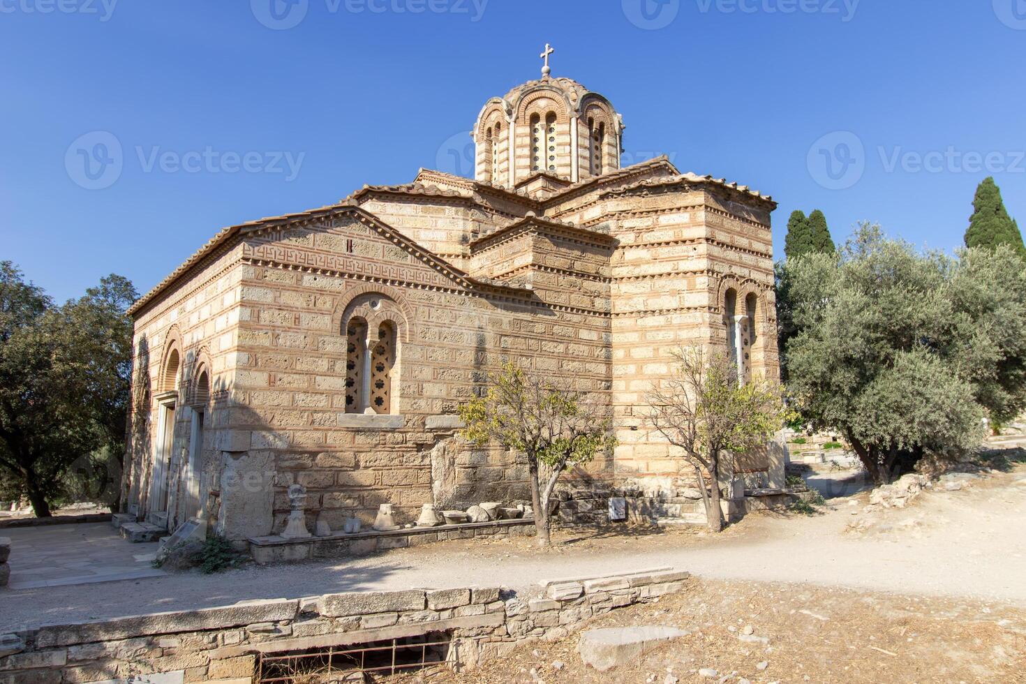 Ancient Byzantine-style stone church with domed roof and cross on top, surrounded by trees and path in Athens, Greece. Outdoor architectural photography photo