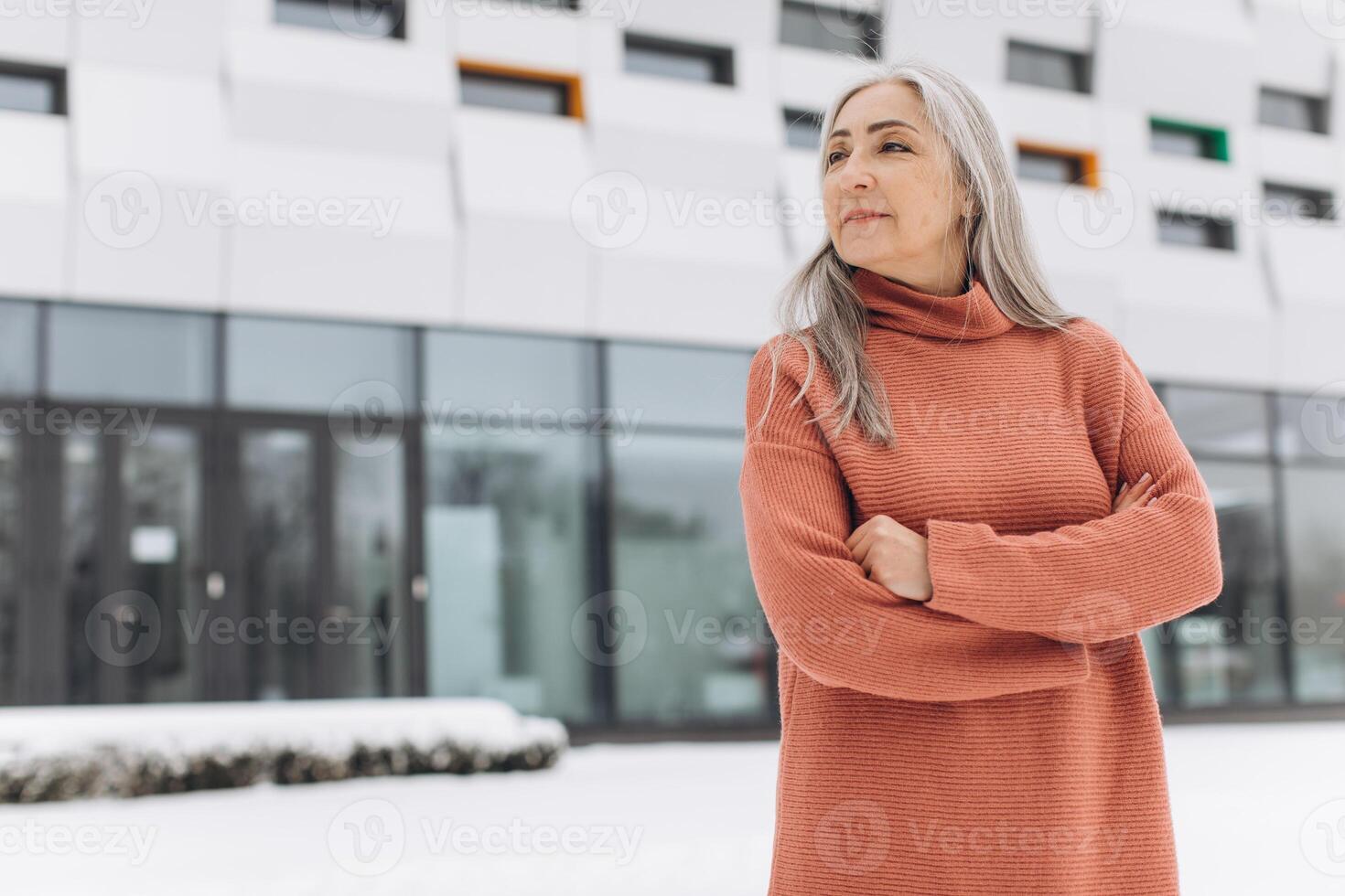 Senior woman with gray hair in a knitted sweater posing on the background of a modern house in winter photo