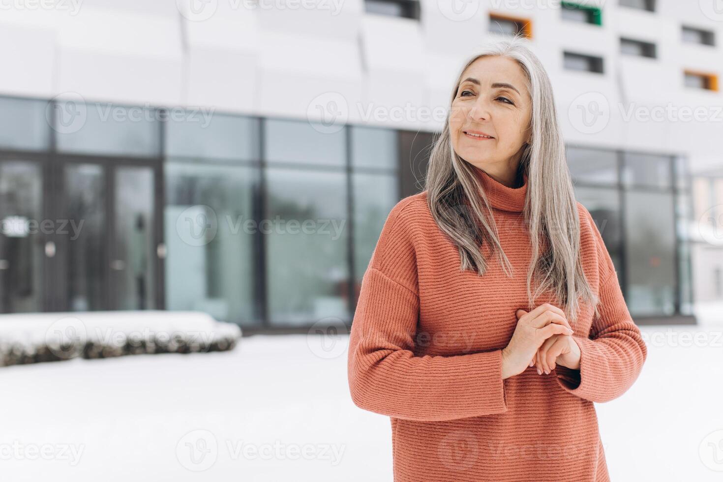 Senior woman with gray hair in a knitted sweater posing on the background of a modern house in winter photo